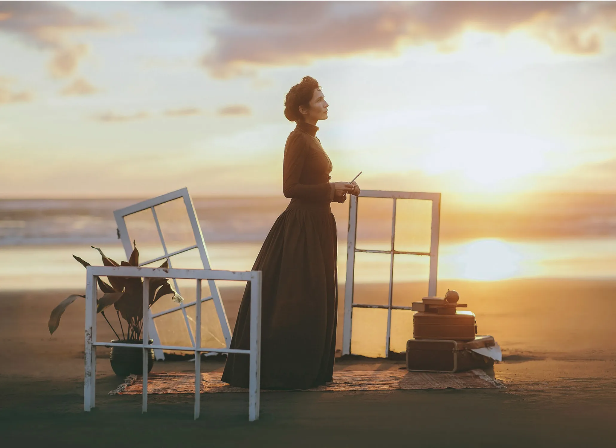 A woman in a dark high-necked period costume standing between two window frames on a beach at sunset